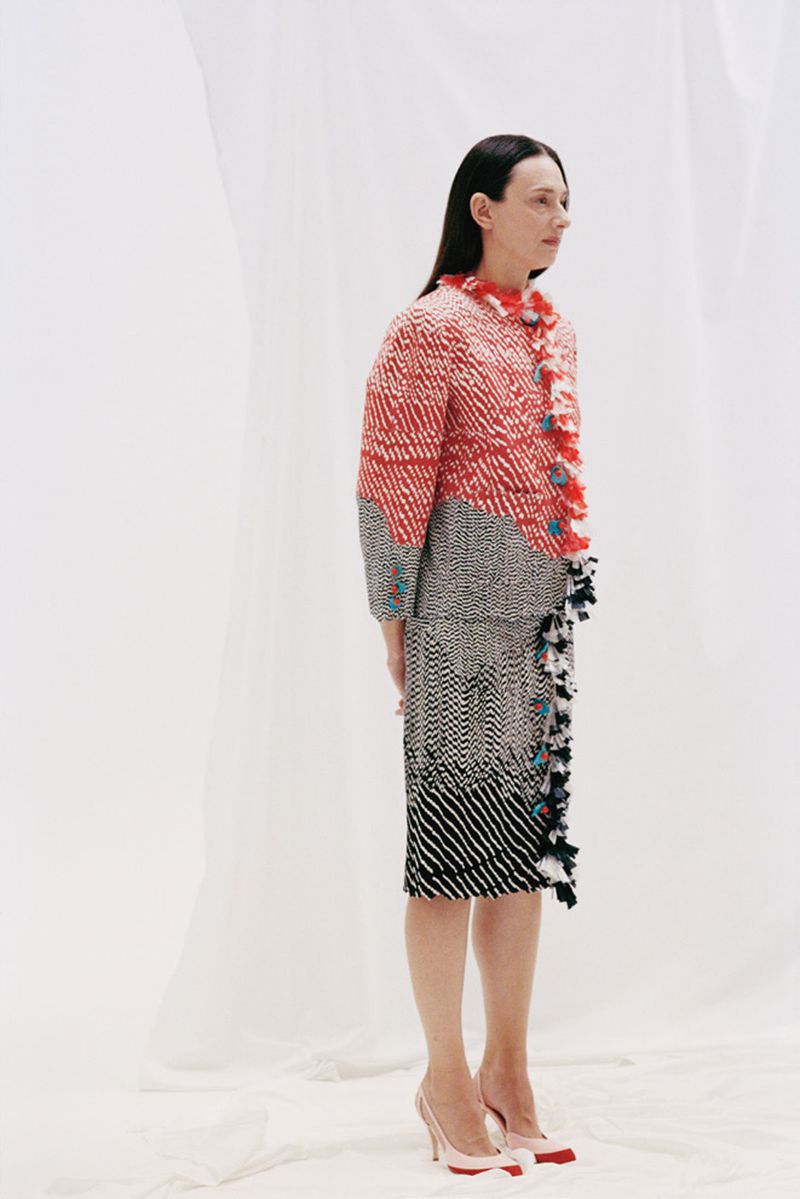 A woman models a red, white, and black patterned jacket and skirt with high heels against a bright white backdrop in a studio setting.
