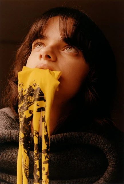 Close-up portrait of a young woman with dark hair and eyes looking up, with a yellow graphic-printed garment held to her mouth.