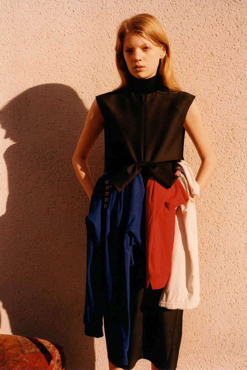 Fashion shot of a blonde woman in a black top and skirt, with colourful shirts attached, against a textured neutral wall with shadow.