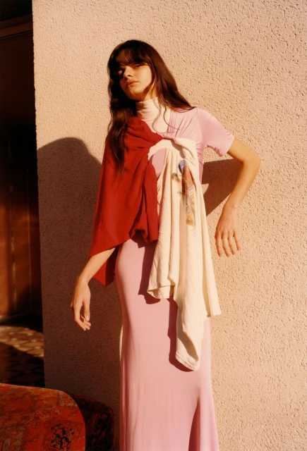 A young woman with long brown hair poses near a textured wall, wearing a pink dress, red shawl, and cream-colored draped garment.