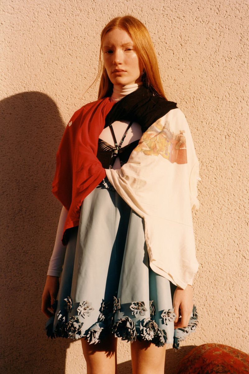 Young woman with long red hair poses in a pastel-blue skirt with flower details and a color-blocked top in front of a peach wall.