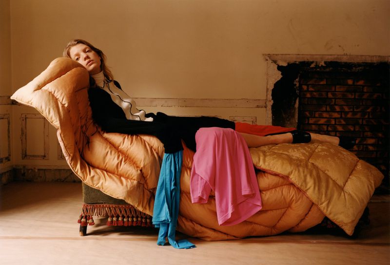 A woman reclines on an antique chaise lounge covered in a padded golden-yellow puffer blanket in a room with a fireplace.