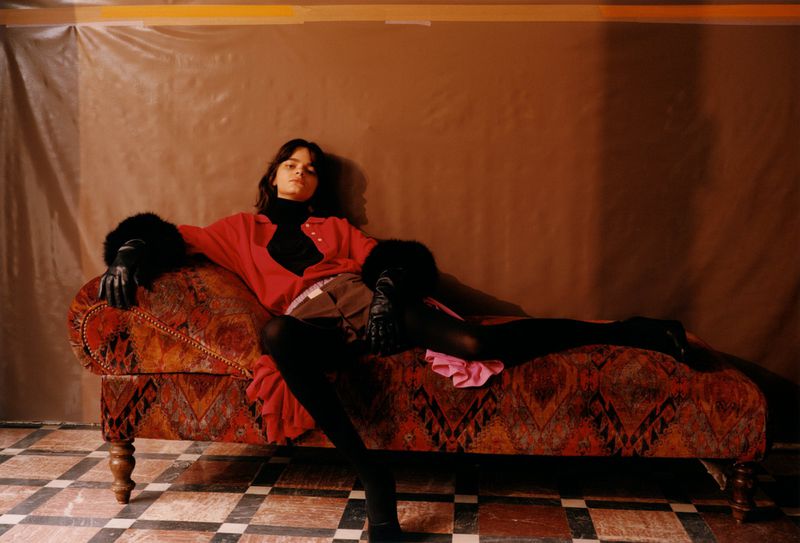 A young adult woman with long brown hair poses on a chaise lounge in a red jacket with black fur cuffs against a brown backdrop.
