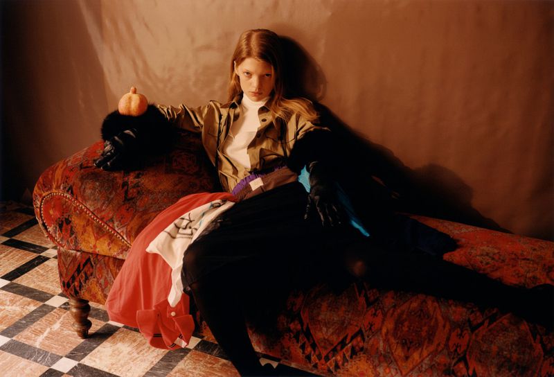 An adult woman poses on an ornate chaise lounge wearing a khaki jacket, black pants, and fur gloves with a pumpkin resting on the gloves.