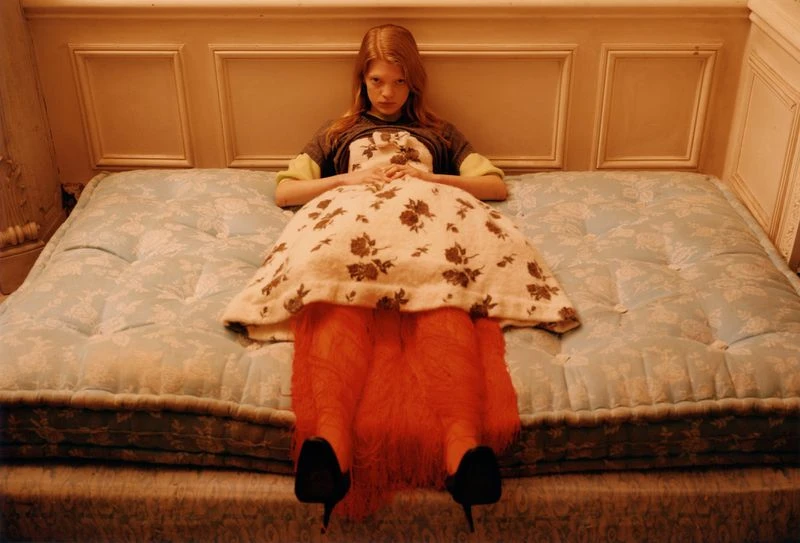 Young woman poses in a patterned dress while lying on a tufted blue mattress indoors in a fashion editorial-style portrait. 