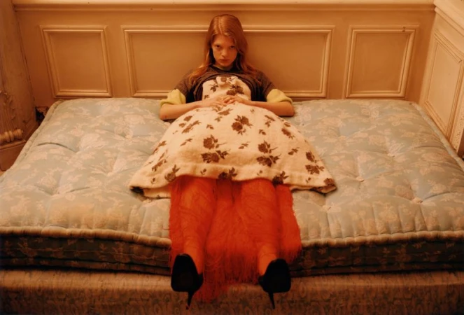 Young woman poses in a patterned dress while lying on a tufted blue mattress indoors in a fashion editorial-style portrait. 