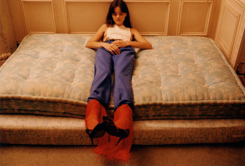 A young woman lies on a floral mattress wearing blue pants and orange heels, casually posed indoors with a neutral expression.