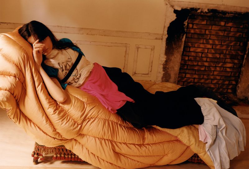 Young adult posing on an oversized quilted chaise lounge in a photo studio setting. Faded colors and relaxed pose creates a unique fashion image.