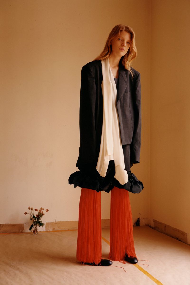 Model standing in an oversized black jacket, white draped top, and long orange fringe pants in an empty warm-toned studio room.