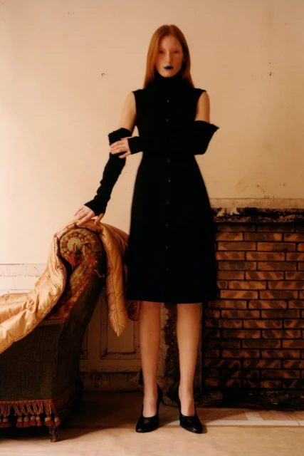 A woman with red hair poses in a black dress and gloves next to a chaise lounge and fireplace in an indoor environment.