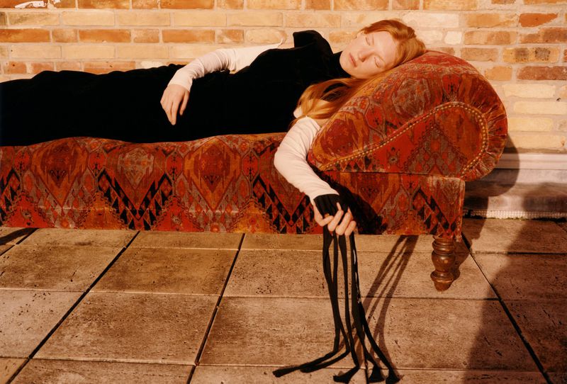 Redhead woman with closed eyes in black dress lies on patterned chaise lounge in front of brick wall, holding black fringe.
