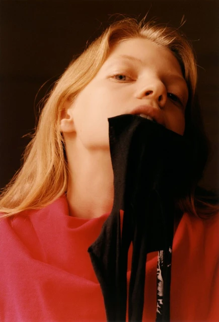 Portrait of a blonde woman with a red shirt, holding black clothing in her mouth in a low-angle shot against a dark background.