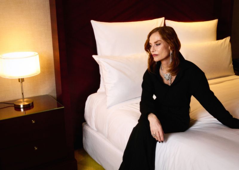 Adult woman in elegant black outfit and statement jewelry poses on a pristine white hotel bed with a warm lamp on a bedside table.