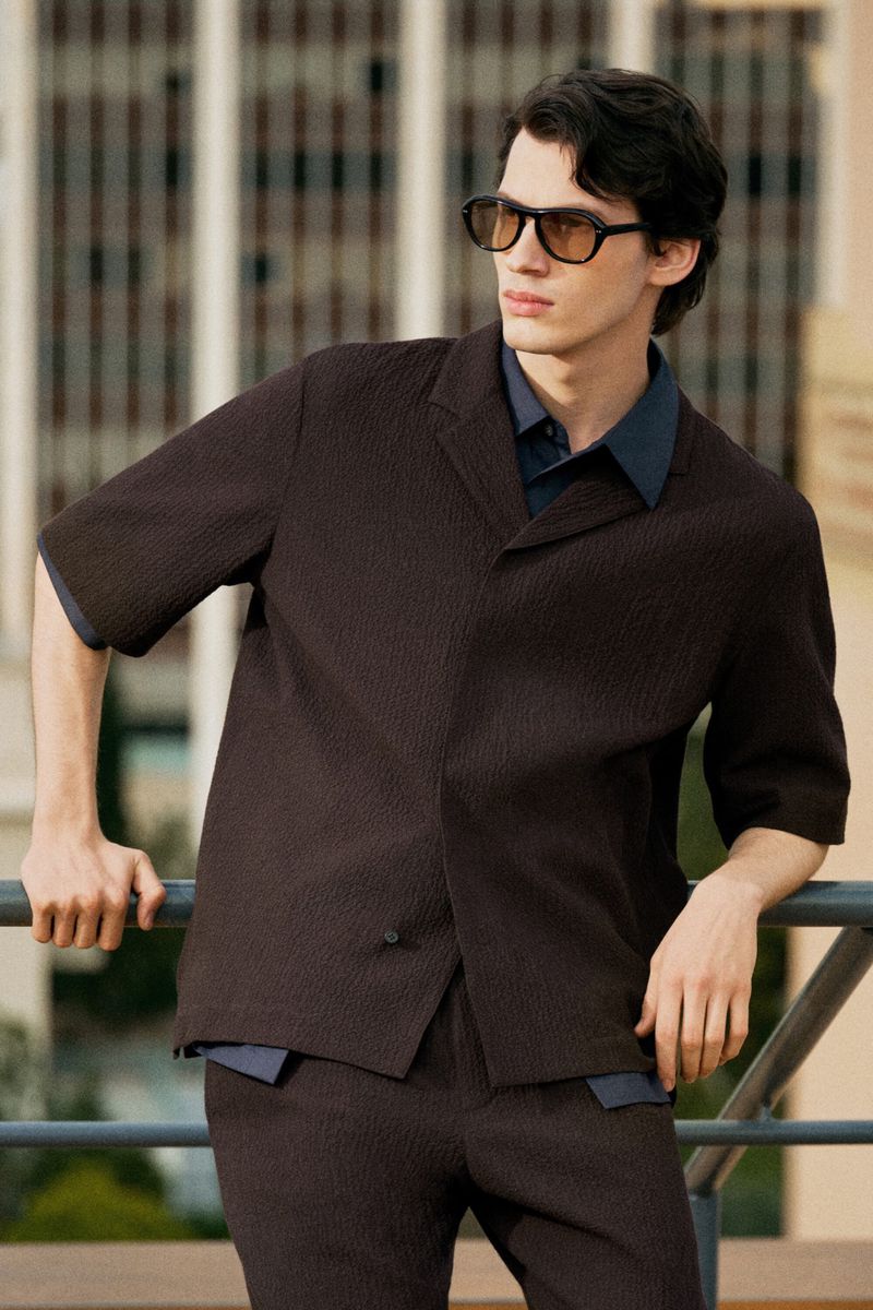 Portrait of a young adult man in a dark brown textured suit and sunglasses, leaning against a metal railing outdoors.