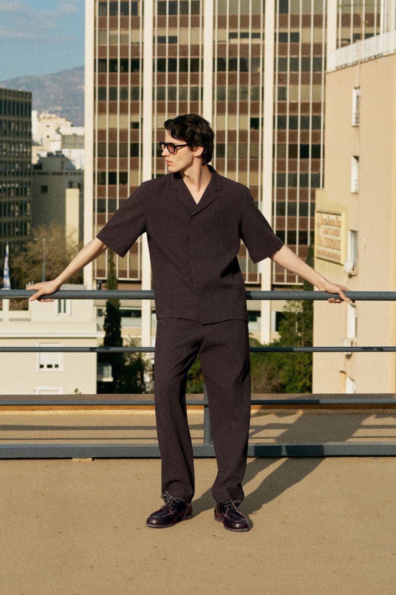 Model Kaplan Hani poses on a rooftop wearing a dark short-sleeve shirt, matching trousers, and sunglasses against a city skyline.