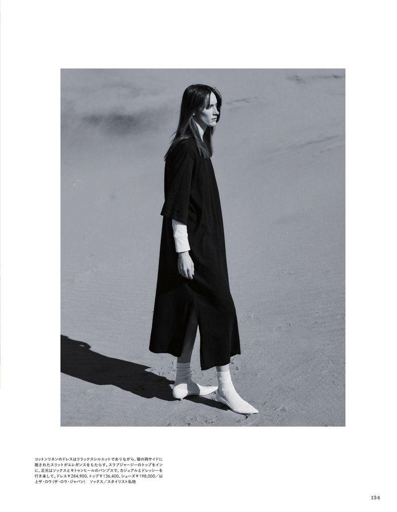 Monochrome fashion shot of a woman in a dark midi dress with long sleeves and white shoes, posing on a light beige sandy surface.