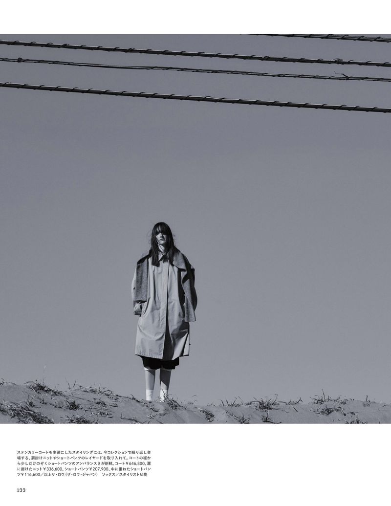 Monochrome full shot of a woman in a trench coat standing on a sand dune beneath telephone wires against a pale, overcast sky.