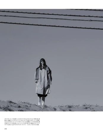 Monochrome full shot of a woman in a trench coat standing on a sand dune beneath telephone wires against a pale, overcast sky.