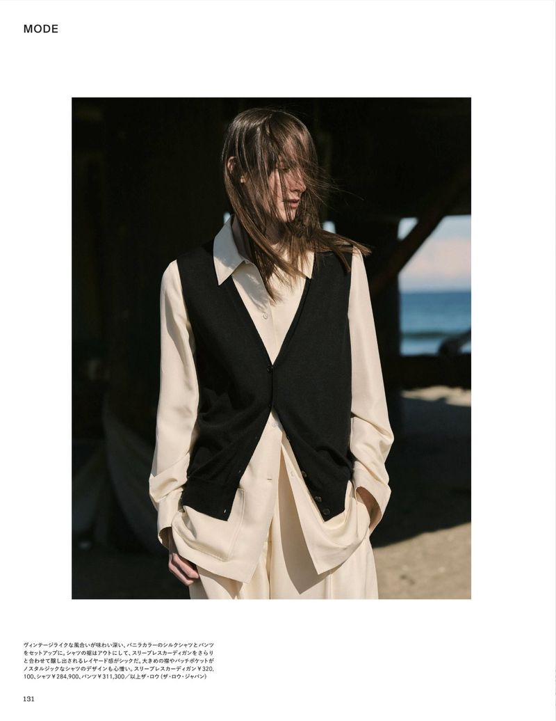 Model wearing a black vest over a cream button-down shirt and trousers standing near a wooden pier on a beach