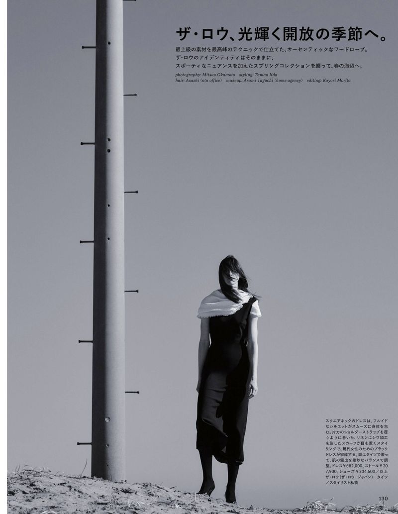 Monochromatic fashion shot of a woman in a black dress and a white scarf, standing on a sandy terrain with a tall pole nearby.