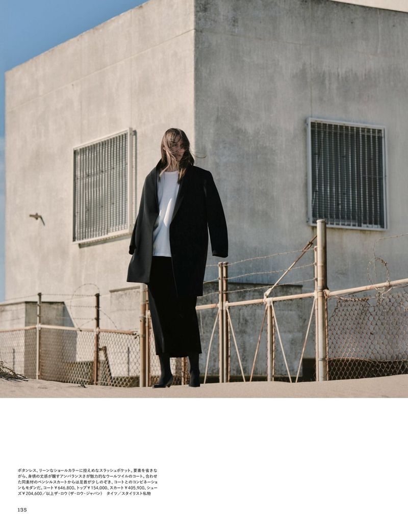 Model Sveta Kare wearing an oversized black coat and white top standing outdoors near a concrete structure with wire fencing.