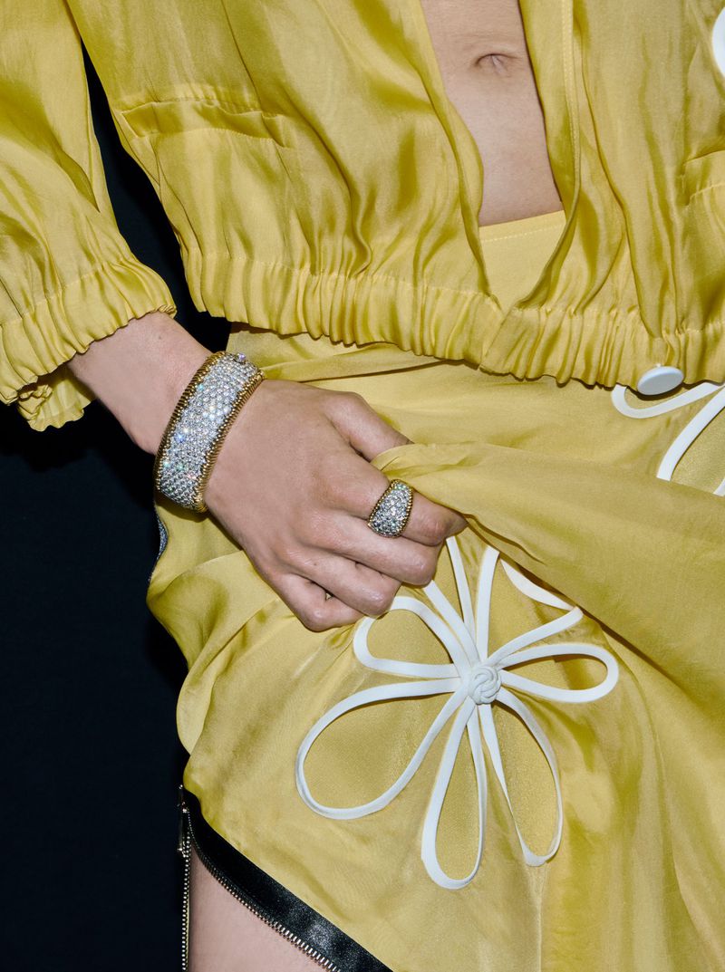 A person in a yellow outfit poses with jewelry. The person wears a matching ring and bracelet, while a white floral design accents the skirt.