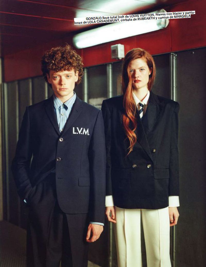 Two fashion models in formal wear stand indoors with serious expressions; one man in LVM suit and one red-haired woman in oversized blazer.