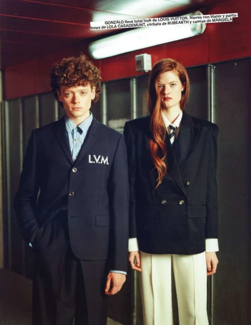 Two fashion models in formal wear stand indoors with serious expressions; one man in LVM suit and one red-haired woman in oversized blazer.