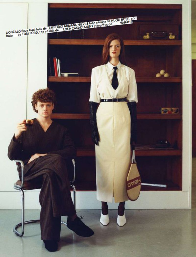 Editorial fashion shot of a man seated with a drink and a woman standing with a tennis racket in an indoor setting.