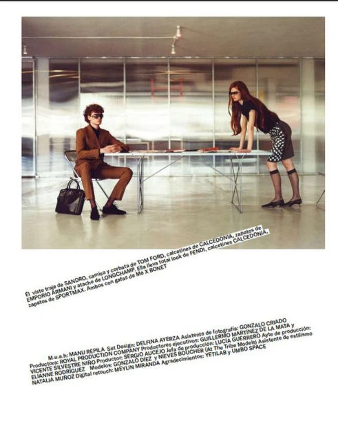 Two fashion models in business attire pose in a modern office. One model leans on a table while the other sits next to a black tote bag.
