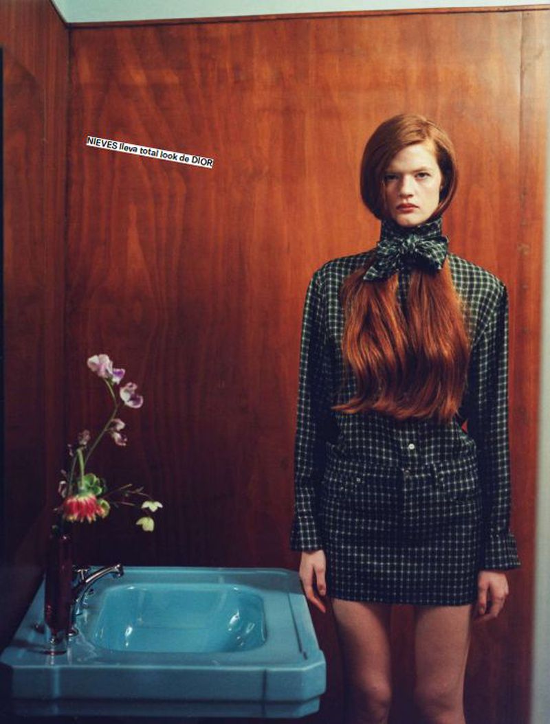 A red-haired woman poses in a plaid outfit with a blue sink in a bathroom setting, capturing a blend of fashion and interior elements.