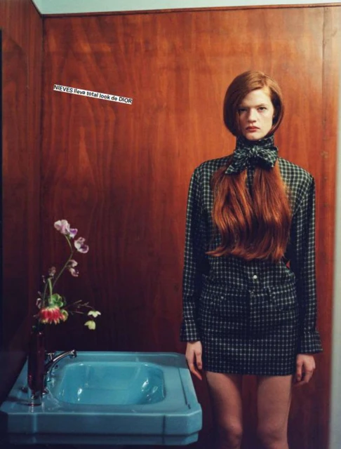 A red-haired woman poses in a plaid outfit with a blue sink in a bathroom setting, capturing a blend of fashion and interior elements.