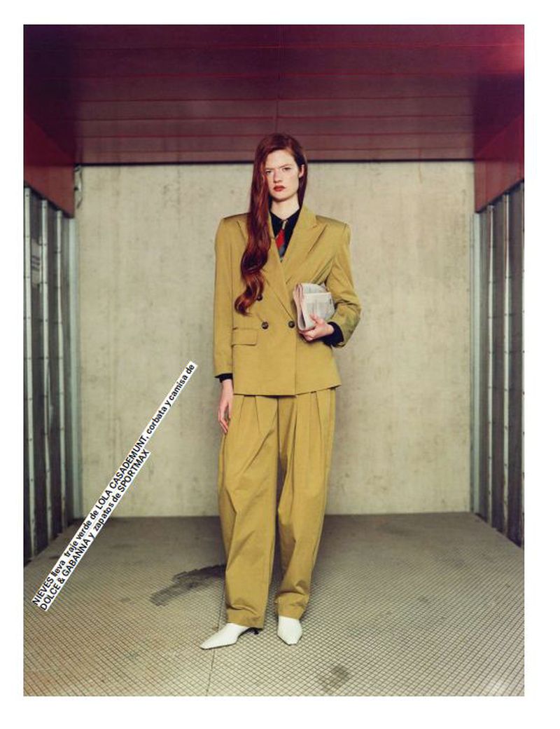 Woman in a mustard-yellow pantsuit poses in an indoor setting. The woman is holding a newspaper with long red hair.