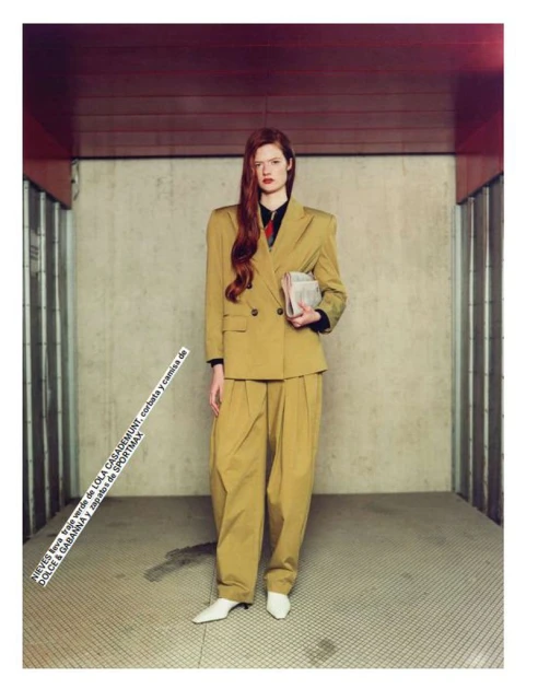 Woman in a mustard-yellow pantsuit poses in an indoor setting. The woman is holding a newspaper with long red hair.