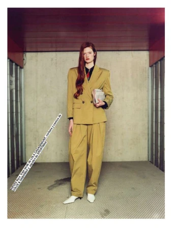Woman in a mustard-yellow pantsuit poses in an indoor setting. The woman is holding a newspaper with long red hair.