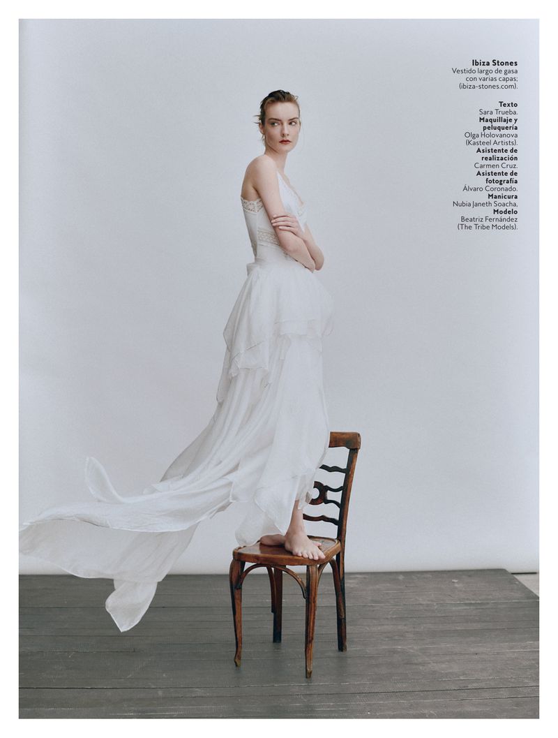 A woman in a white dress poses while standing on a wooden chair in front of a white background in a studio with natural light.