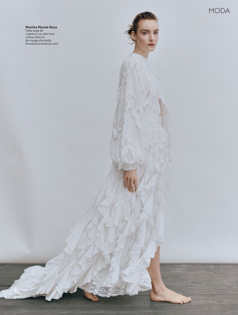 A woman is elegantly walking while wearing a long white dress with ruffles, posing against a neutral studio backdrop in a fashion editorial shot.