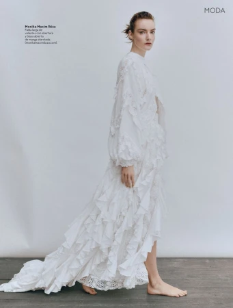 A woman is elegantly walking while wearing a long white dress with ruffles, posing against a neutral studio backdrop in a fashion editorial shot.