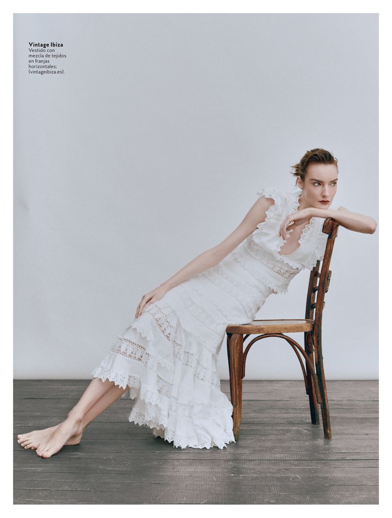 Woman in a white lace dress poses while seated on a wooden chair against a white background in a fashion editorial style image.