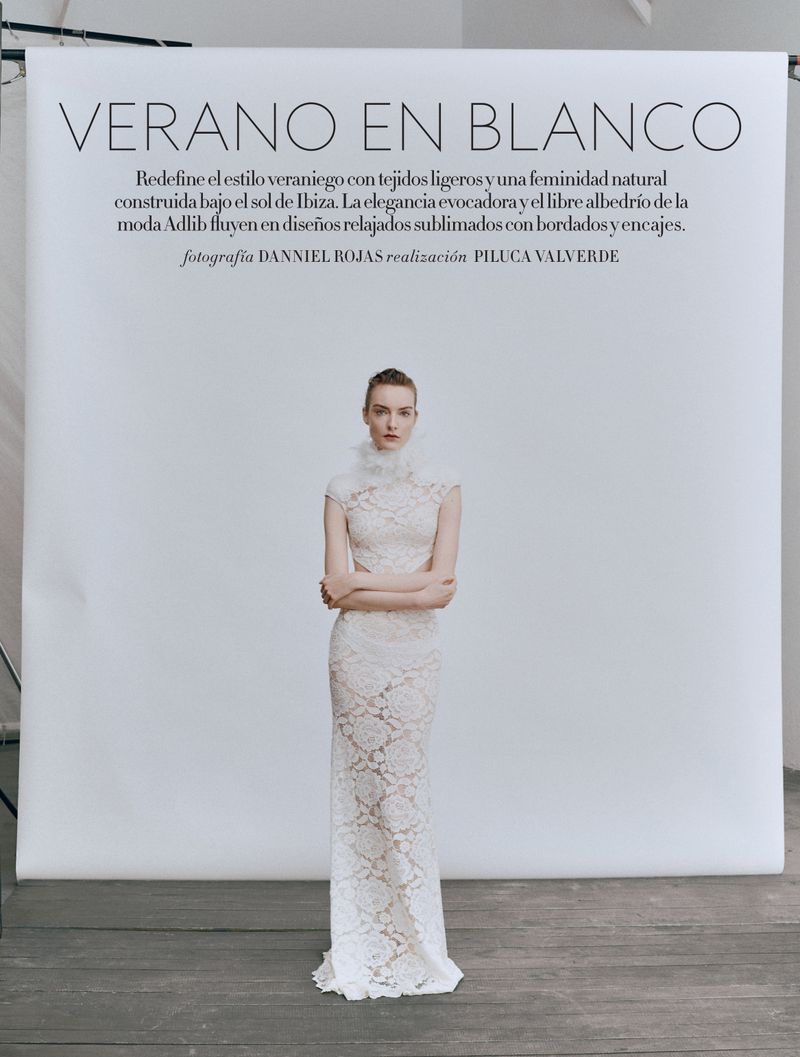 Fashion editorial shot of a slim, adult female model wearing a white lace gown with a high ruffle collar posing against a white backdrop.