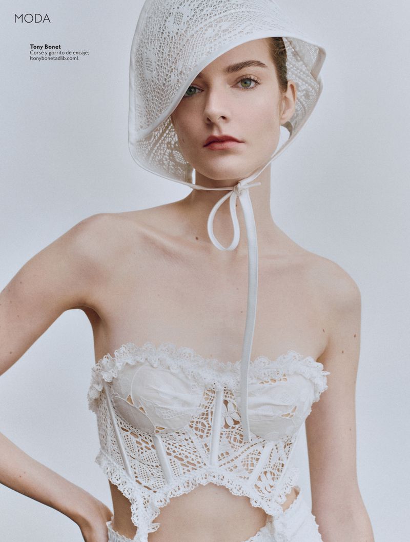 A fair-skinned adult female poses in a white, lacy lingerie top and matching hat in a studio with natural light. Fashion editorial.
