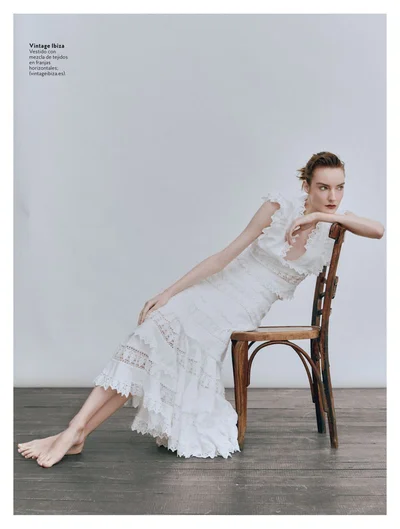 Model Beatriz Fernandes barefoot in a tiered white lace dress seated on a wooden chair against a light studio backdrop.