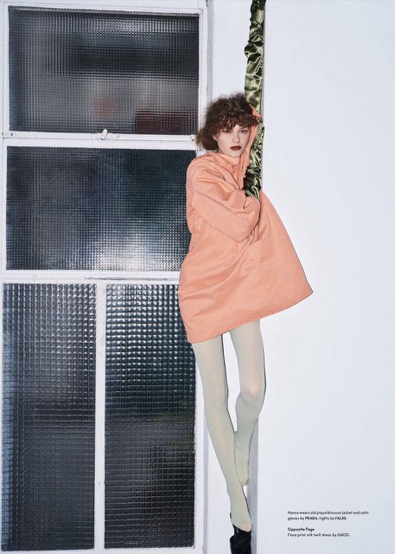 Fashion photo of a slender adult woman with curly hair, wearing a peach-colored coat, light-green tights, and black heels near a window.