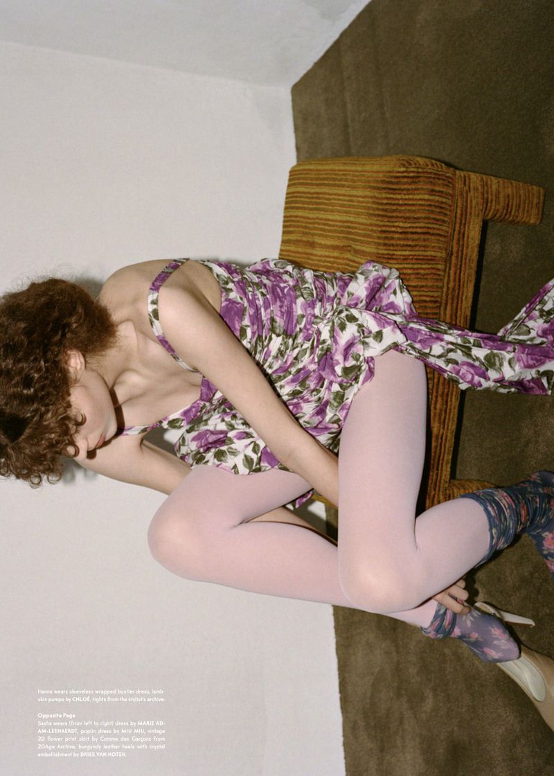 Woman in floral dress with tights and heels poses on a chair in an indoor setting, creating a fashion editorial shot.
