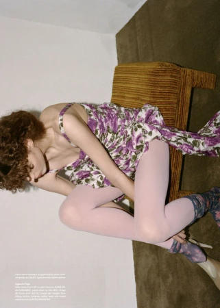Woman in floral dress with tights and heels poses on a chair in an indoor setting, creating a fashion editorial shot.