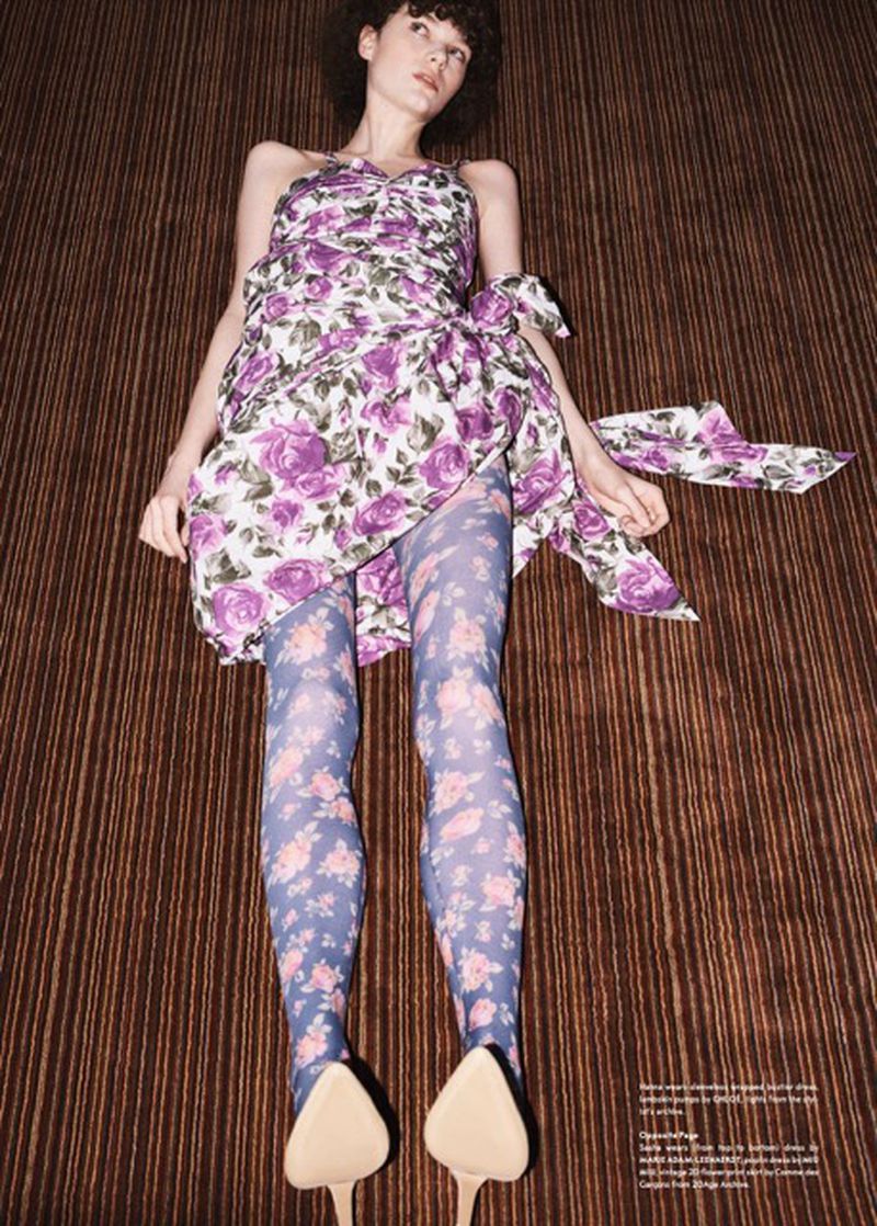 Woman in floral mini dress and tights poses on brown striped surface. She is lying down, looking left with dark curly hair and light skin.