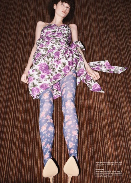 Woman in floral mini dress and tights poses on brown striped surface. She is lying down, looking left with dark curly hair and light skin.