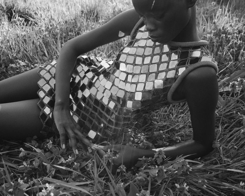 Woman in a mirrored dress lies in the grass with wildflowers. Black and white fashion portrait taken outdoors.