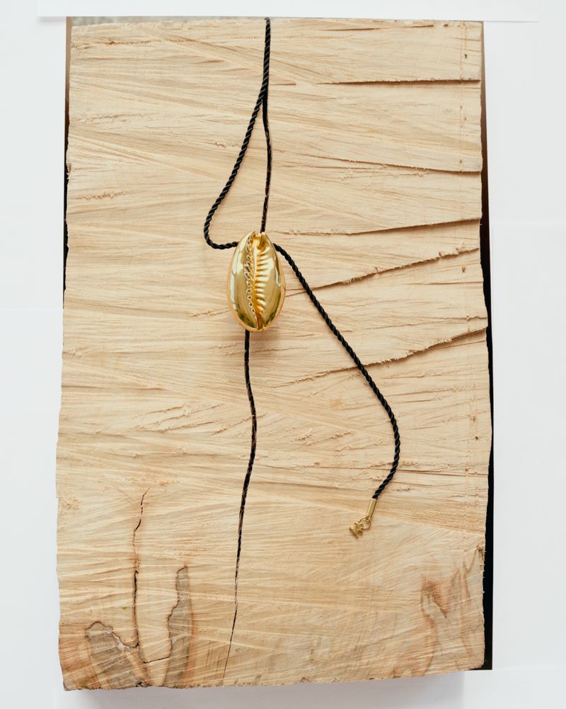 Still life of gold cowrie shell pendant on black cord necklace, displayed against a light wood grain background and white surface.
