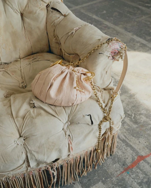 Light pink purse with a gold chain rests on a distressed off-white armchair with fringe detailing against a grey studio backdrop.